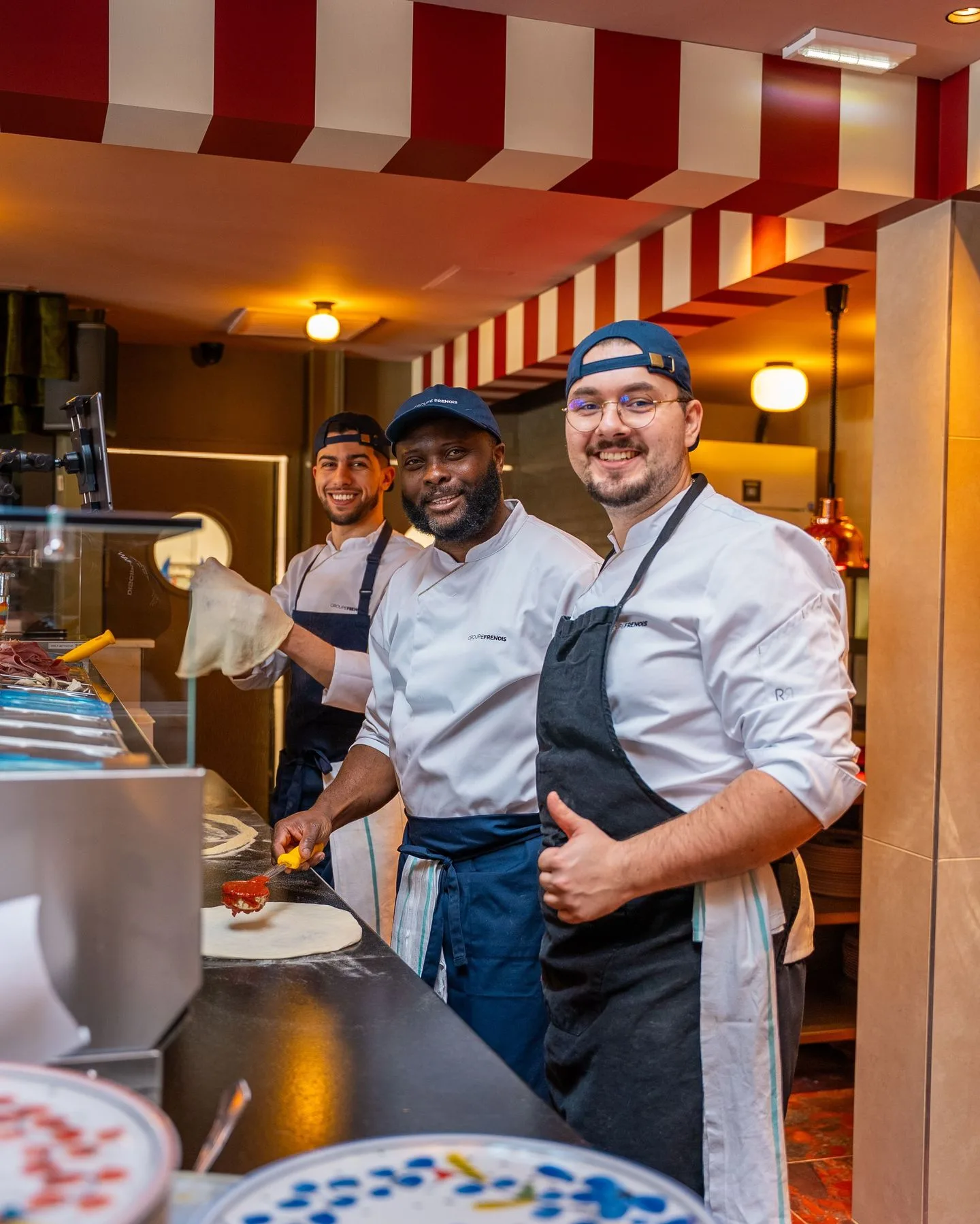 Pizzaiolos de l'équipe Carla en cuisine, en train de garnir une pizza, sous un dais rayé rouge et blanc