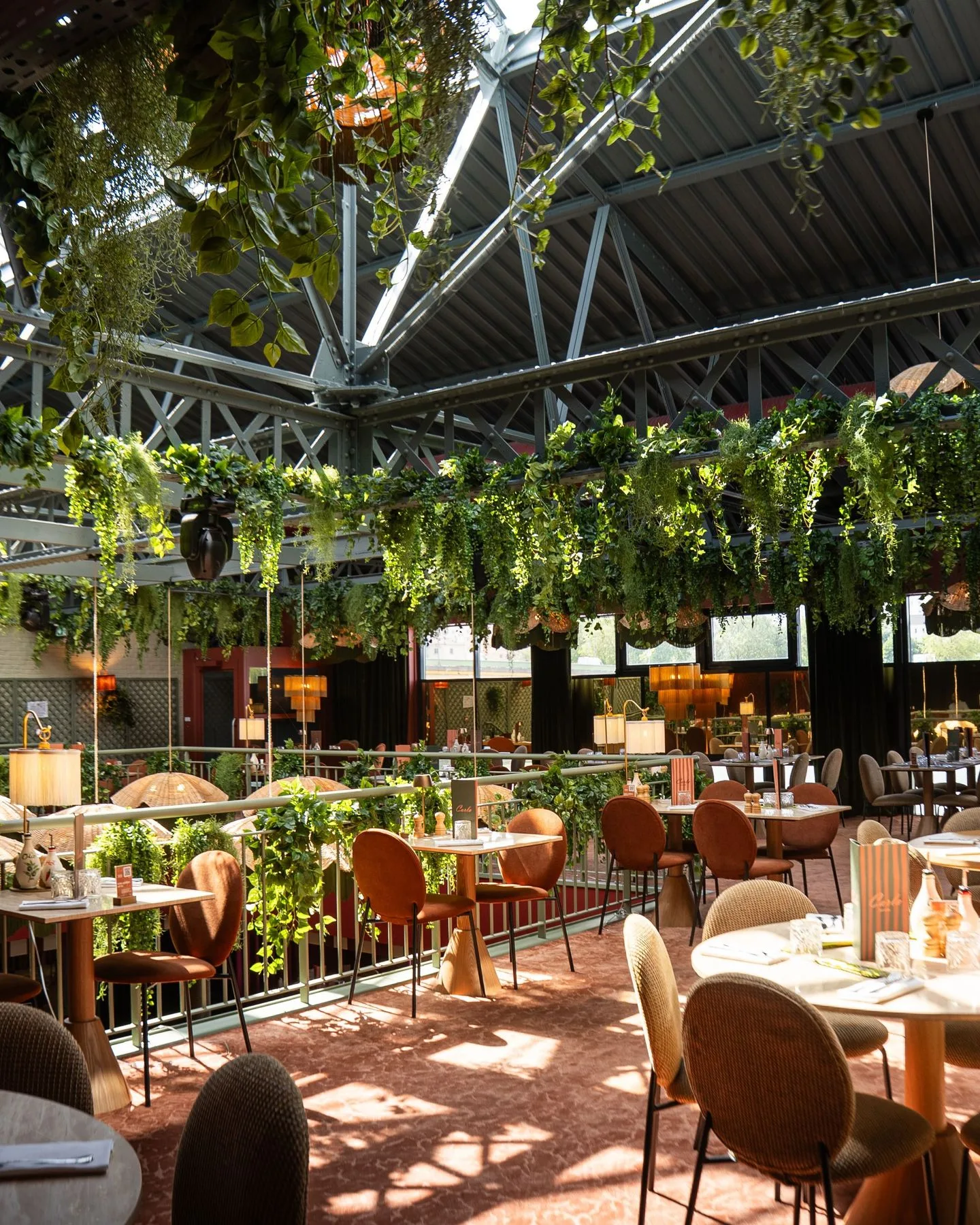 Salle du restaurant Carla sous une verrière industrielle, lianes de plantes vertes en suspension, chaises terracotta et sol carrelé terracotta