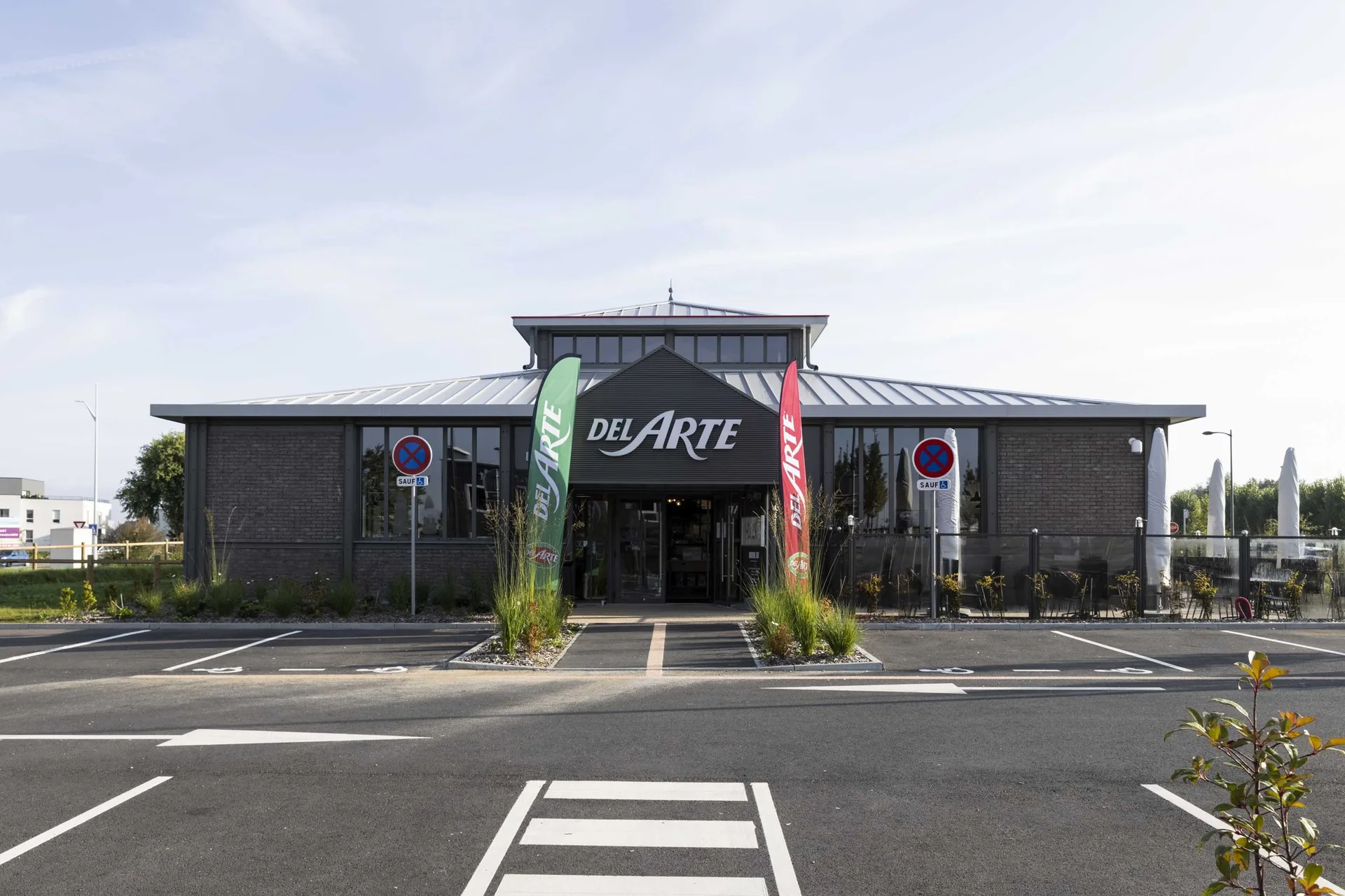 Vue frontale du Del Arte Isneauville depuis le parking, avec deux beach flags vert et rouge Del Arte encadrant l'entrée principale surmontée du logo de l'enseigne, terrasse aperçue à droite