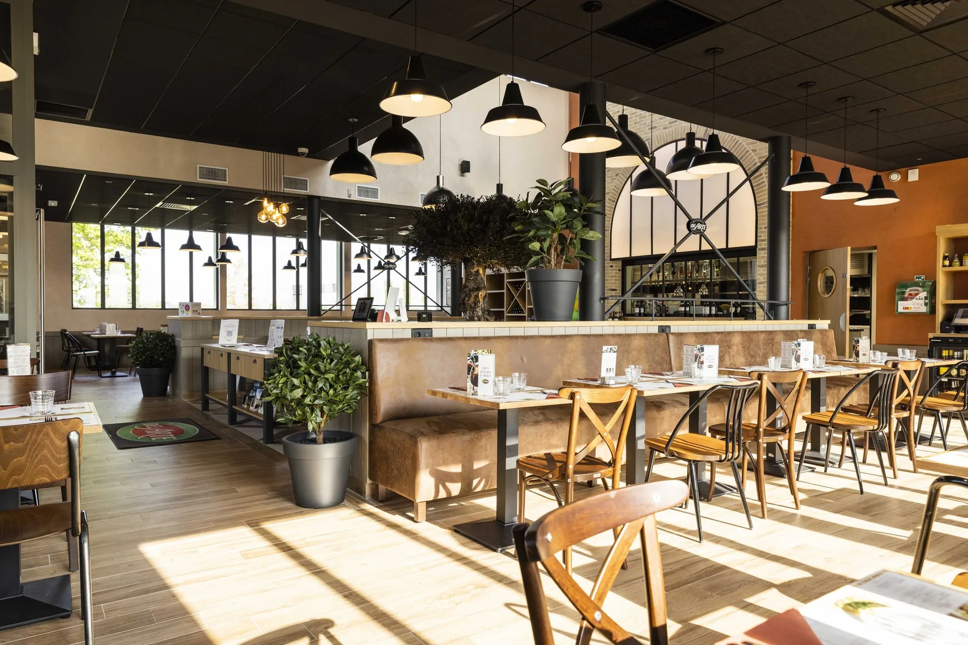 Salle intérieure du Del Arte Isneauville avec banquettes en cuir clair, tables en bois et chaises bistrot, luminaires noirs suspendus, plantes vertes et touches de mur orange, ambiance trattoria italienne moderne