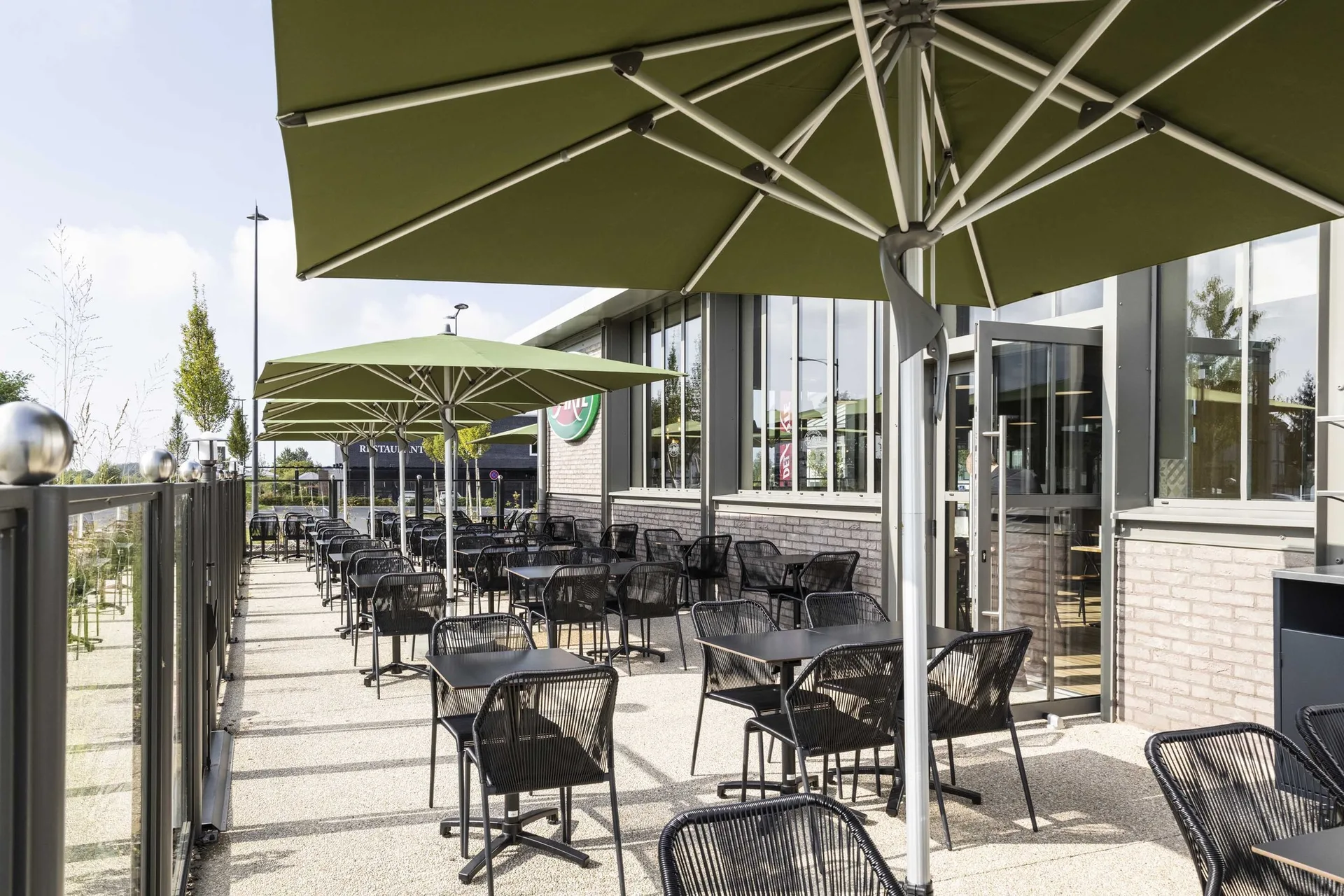 Terrasse extérieure du Del Arte Isneauville avec grands parasols vert olive, tables et chaises noires métalliques le long de la façade vitrée du restaurant