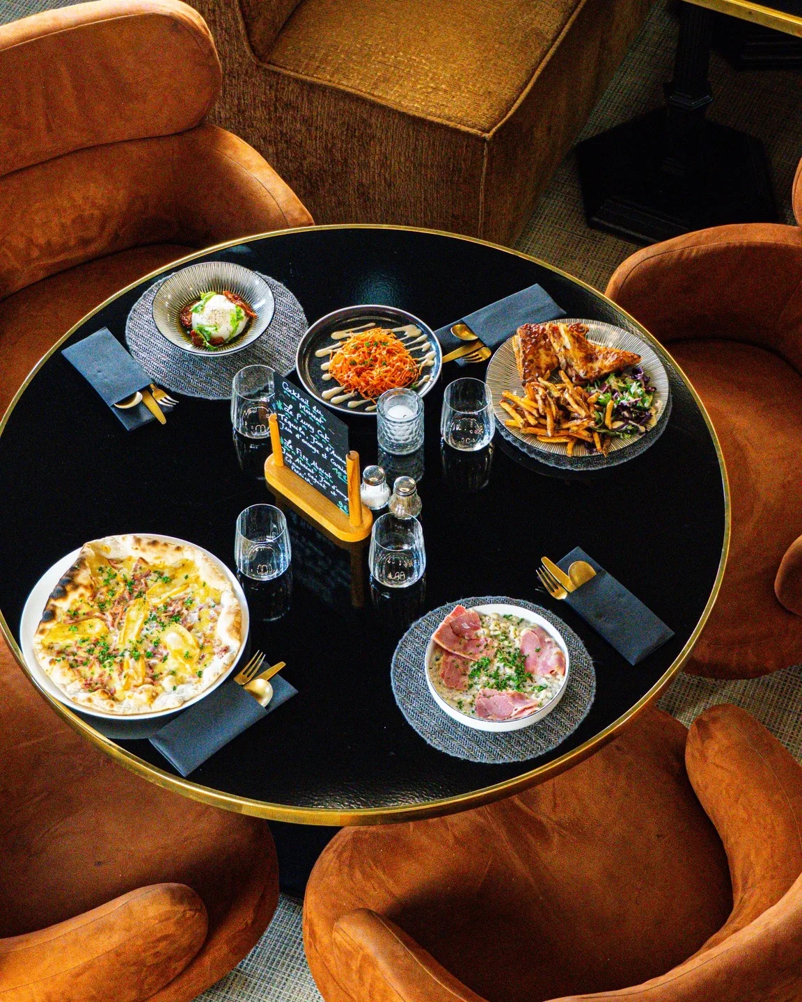 Table ronde noire vue du dessus avec une sélection de plats du Bouillon d'Isno : pizza façon flambée, salade verte à l'œuf, tartare et plat de viande accompagné de frites.
