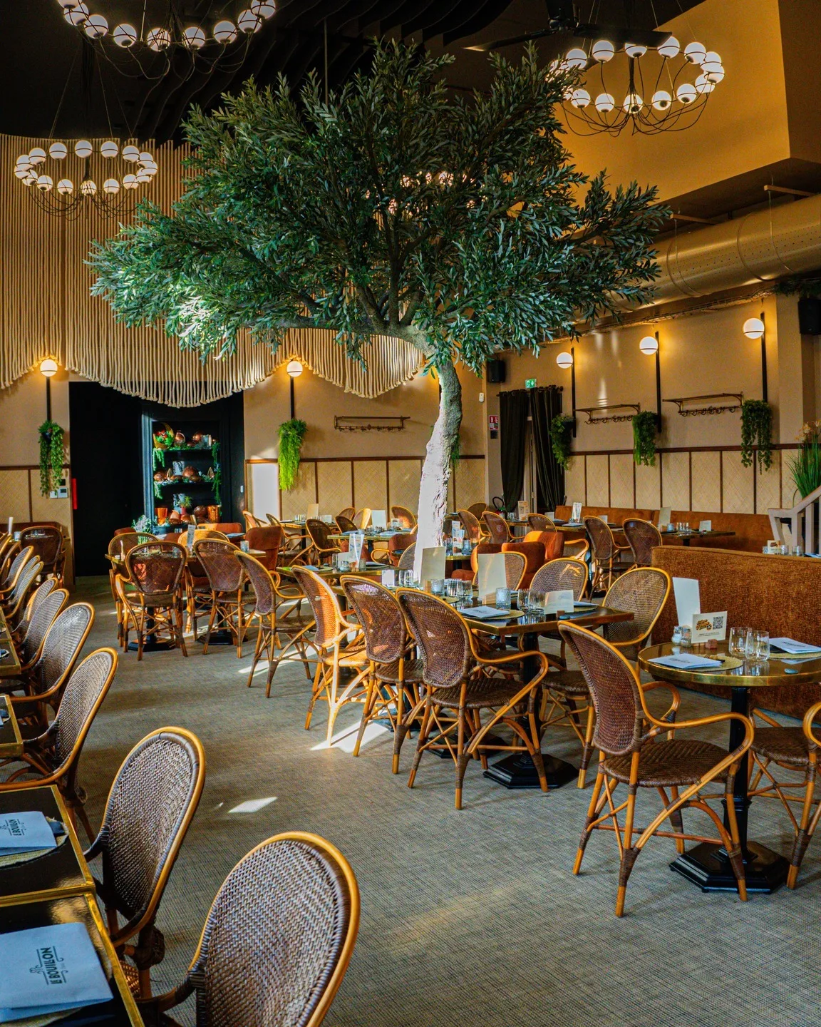 Salle du restaurant à l'ambiance feutrée avec un grand arbre décoratif suspendu, lustres globes dorés et chaises bistrot en rotin autour de tables sombres.
