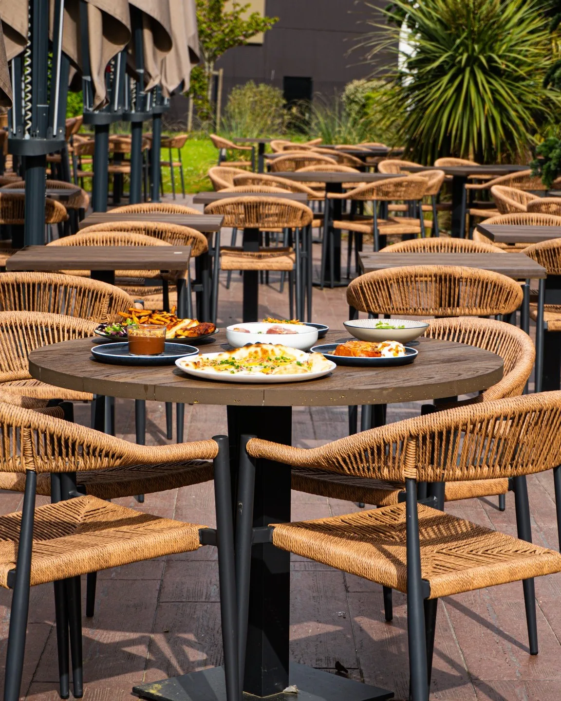 Terrasse extérieure du Bouillon d'Isno avec table ronde dressée de plats à partager au centre, chaises en rotin tressé et parasols repliés en arrière-plan.