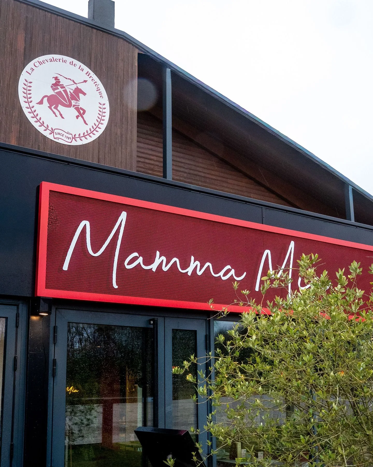 Enseigne Mamma Mia en néon rouge sur la façade en bois du restaurant, sous le logo La Chevalerie de la Bretèque