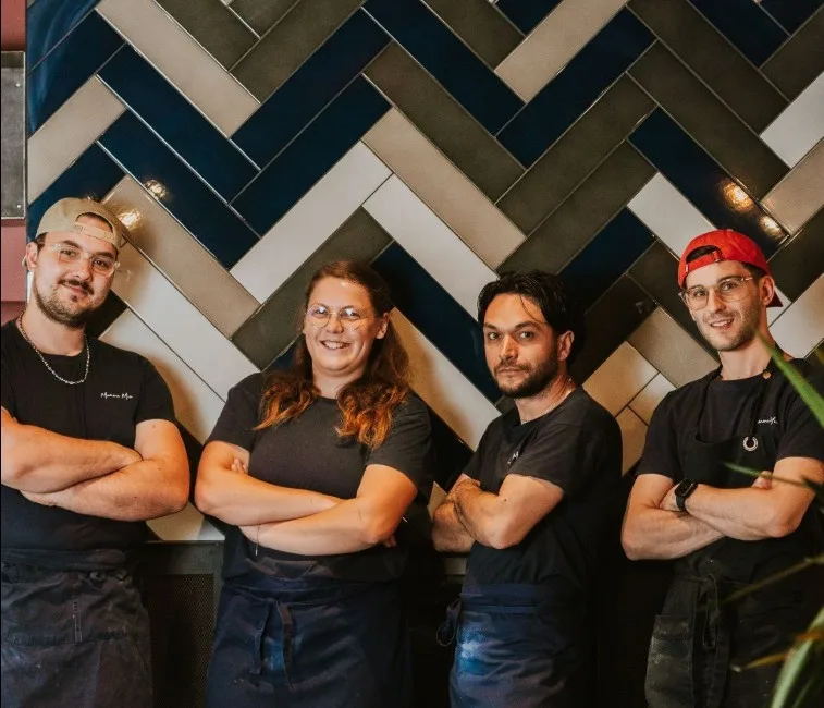Équipe de salle et de cuisine du restaurant Mamma Mia en t-shirts noirs, devant un mur en mosaïque chevrons gris et bleu