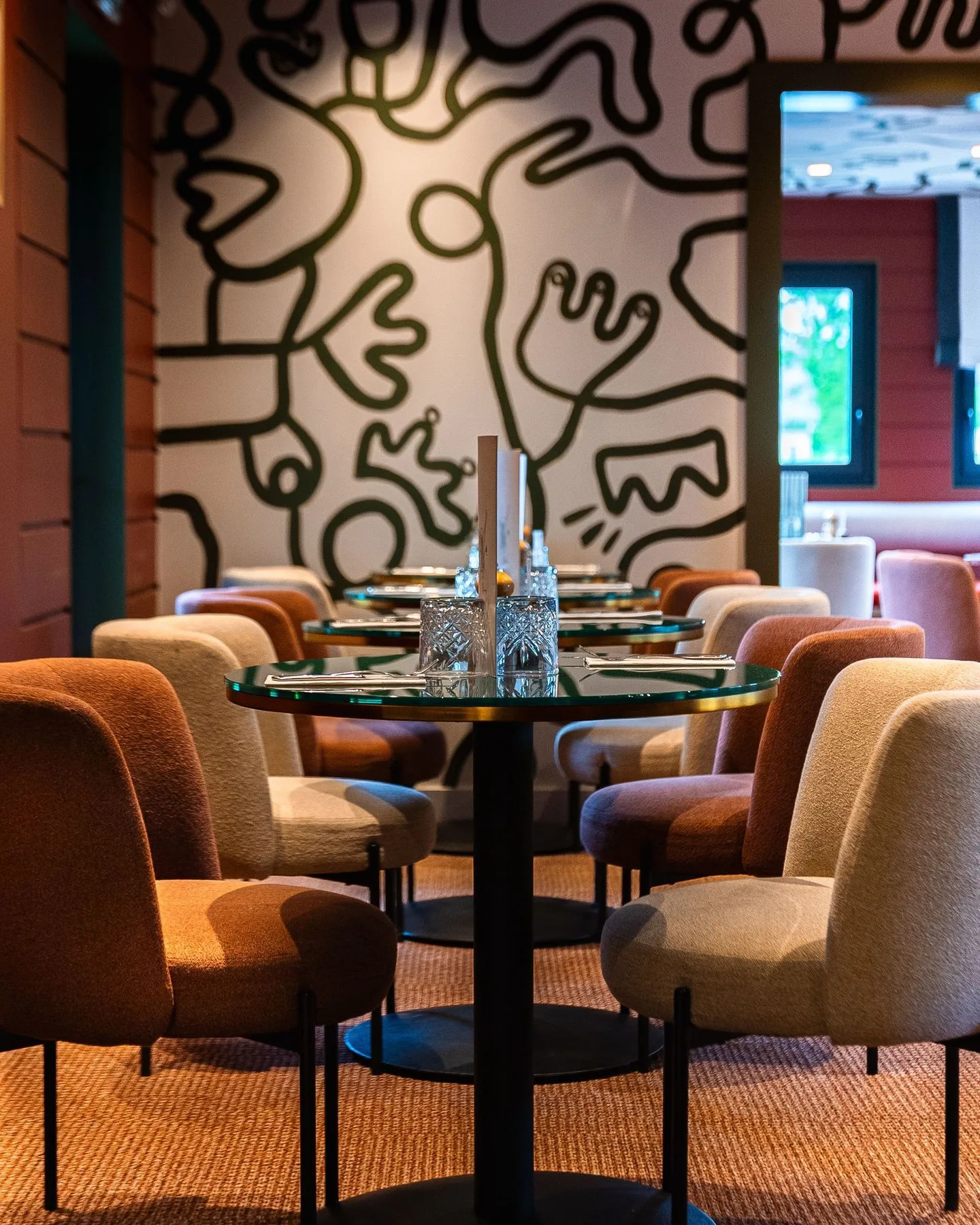 Salle du restaurant Mimosa : tables rondes, chaises en velours terracotta et beige, fresque graphique noir et blanc en arrière-plan