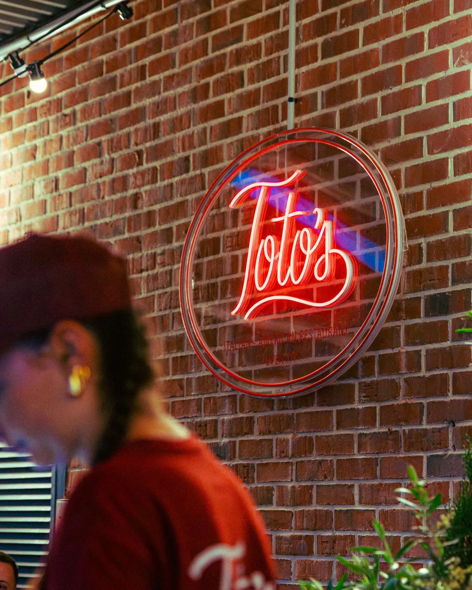 Enseigne lumineuse rouge Toto's sur un mur de briques, avec une membre de l'équipe en t-shirt Toto's au premier plan.
